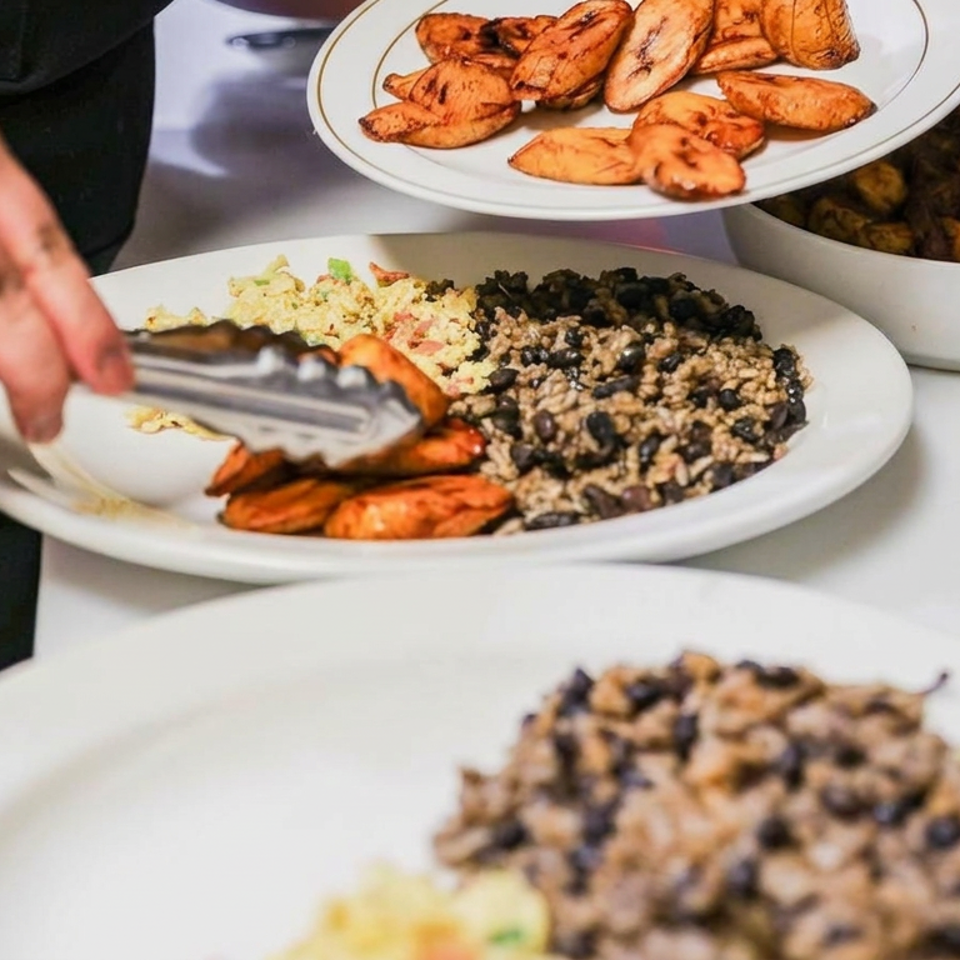 Server uses tongs to plate roasted sweet potatoes alongside black beans, rice, and vegetables at a buffet.